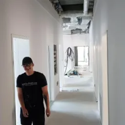 Korytarz biurowy w trakcie odświeżania, widoczne świeżo pomalowane ściany w jasnym odcieniu, instalacje pod sufitem oraz uśmiechnięty malarz w czarnej koszulce z logo firmy.