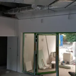 Wnętrze biura w trakcie remontu: ściany po szpachlowaniu, widoczne instalacje pod sufitem, pracownik w tle.