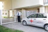 Mężczyzna w białej koszuli i krawacie otwiera białą bramę wjazdową, obok zaparkowany srebrny samochód z logo firmy ochroniarskiej i napisem 'CITY SECURITY'.