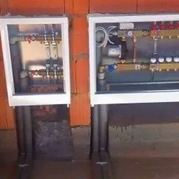 Dwa rozdzielacze ogrzewania podłogowego w białych obudowach, zamontowane na ścianie z czerwonej cegły, z widocznymi rurami doprowadzającymi.