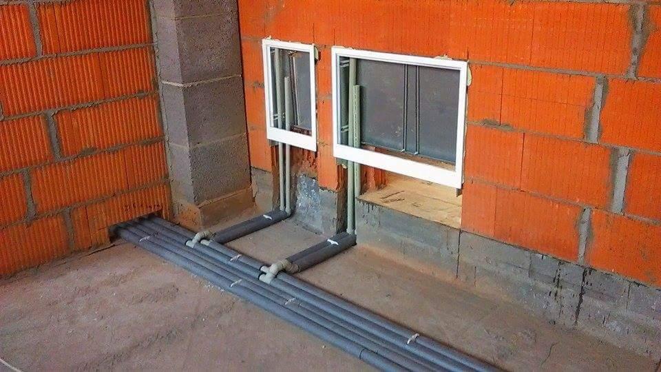 Szare rury instalacyjne prowadzone wzdłuż podłogi i w ścianie z czerwonej cegły, z dwoma białymi ramkami otworów inspekcyjnych w ścianie.