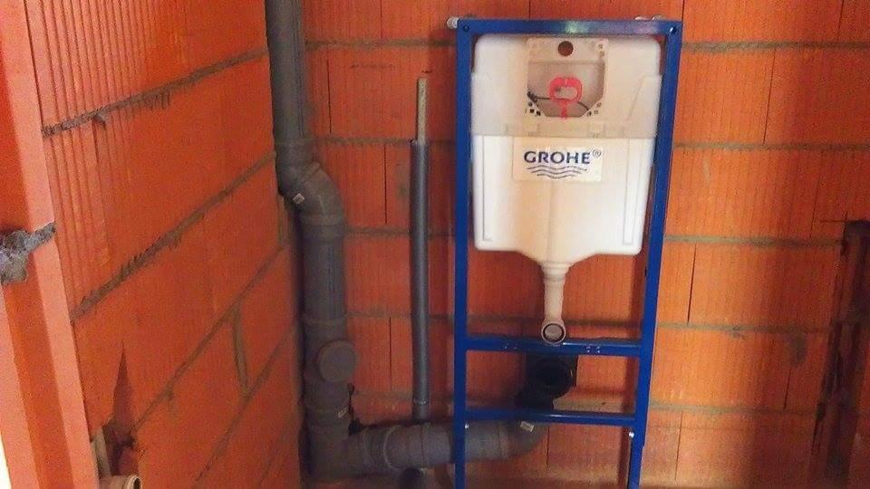 Stelaż podtynkowy Grohe zamontowany na tle surowej ściany z cegły, widoczne szare rury kanalizacyjne.