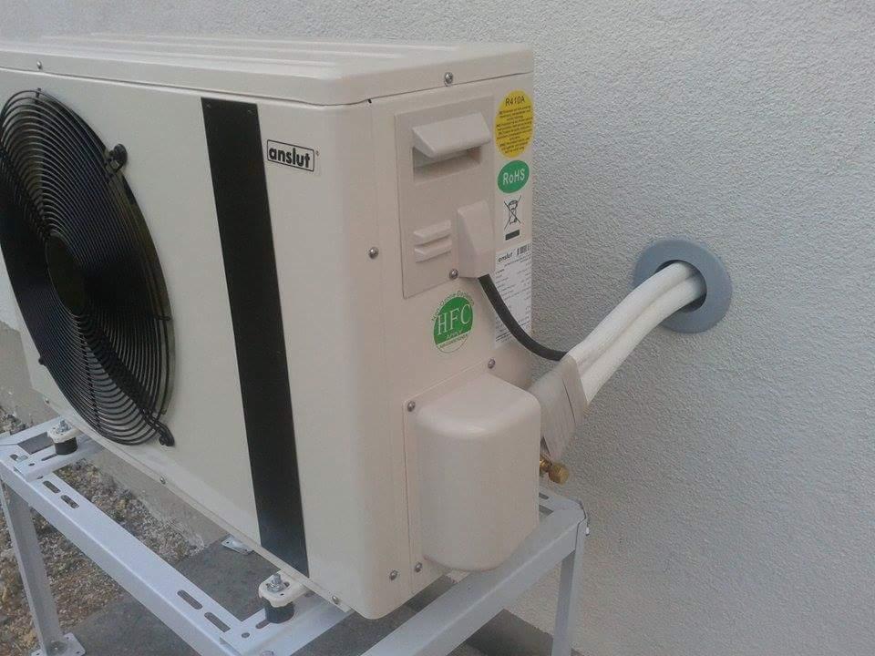 Zamontowana zewnętrzna jednostka pompy ciepła marki Anslut na metalowym stelażu, z widocznymi przewodami chłodniczymi przechodzącymi przez otwór w szarej ścianie.