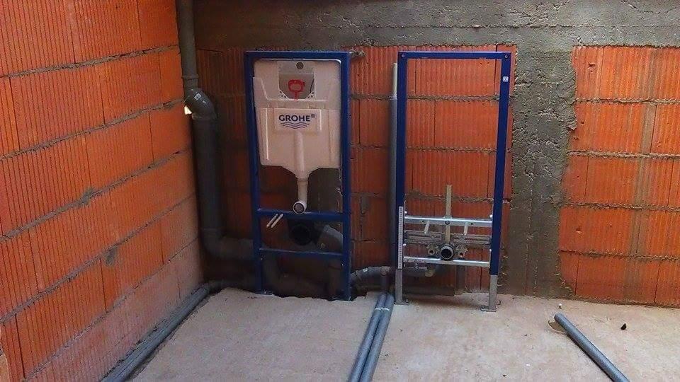 Stelaż podtynkowy WC marki Grohe, z widocznymi rurami kanalizacyjnymi i doprowadzającymi wodę, zamontowany w surowym wnętrzu z cegły i betonu.