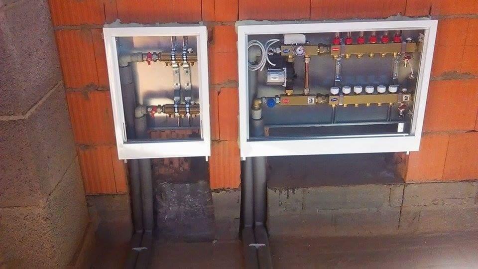 Dwa rozdzielacze ogrzewania podłogowego w białych obudowach, zamontowane na ścianie z czerwonej cegły, z widocznymi rurami doprowadzającymi.