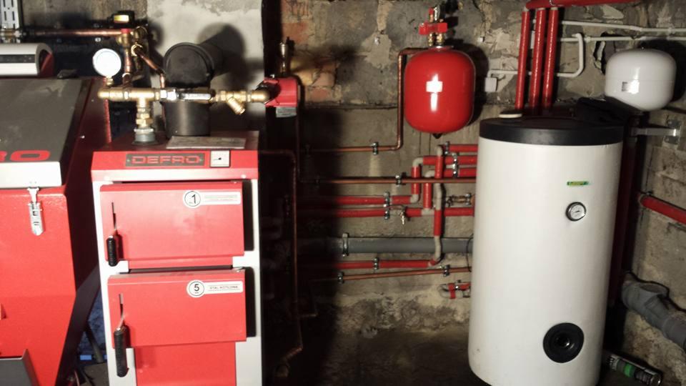 Czerwony piec CO marki Defro z podłączonymi miedzianymi rurami i zbiornikiem akumulacyjnym w kotłowni o surowych ścianach z cegły i betonu.