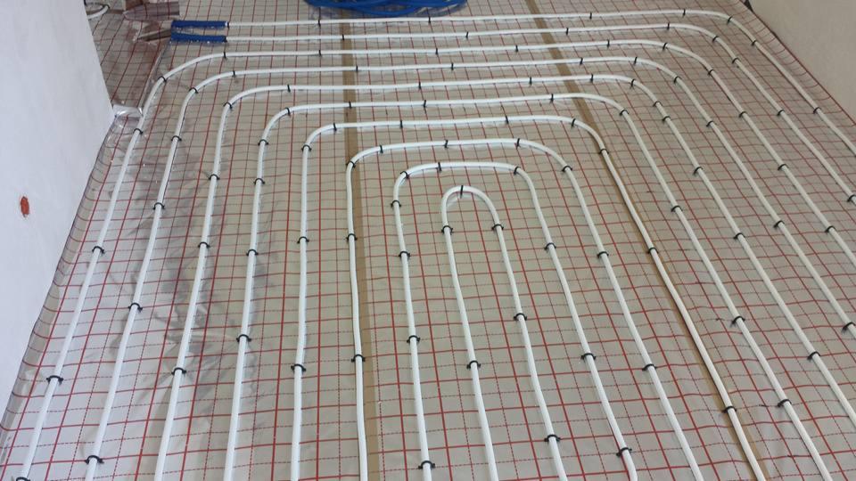 Białe rury ogrzewania podłogowego ułożone w pętle na izolacji z siatką w kolorze czerwono-białym, zamocowane czarnymi uchwytami.