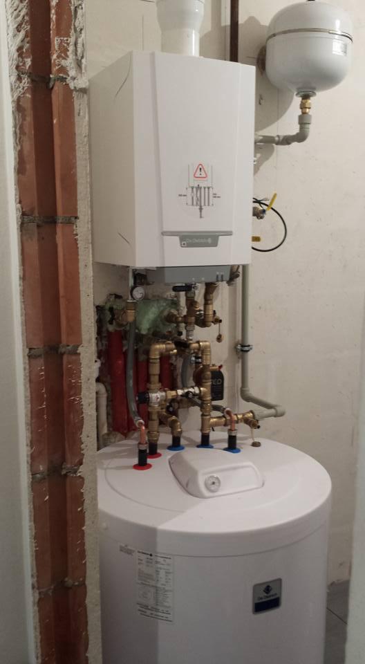 Biały piec centralnego ogrzewania zamontowany nad białym zasobnikiem ciepłej wody, widoczne rury miedziane i zawory, fragment ściany z odkrytą cegłą.