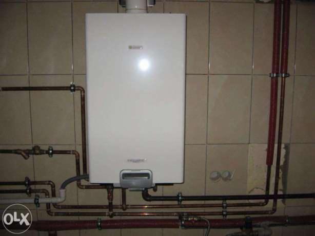 Biały piec gazowy zamontowany na kafelkowanej ścianie z widocznymi rurami miedzianymi i elementami instalacji, w tym fragmentem szarej rury odprowadzającej spaliny.