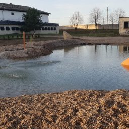 Tomo Bud - Nowo założony staw z fontanną i dekoracyjną amforą, otoczony kamieniami, w tle budynki gospodarcze i drzewa.