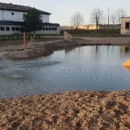 Nowo założony staw z fontanną i dekoracyjną amforą, otoczony kamieniami, w tle budynki gospodarcze i drzewa.