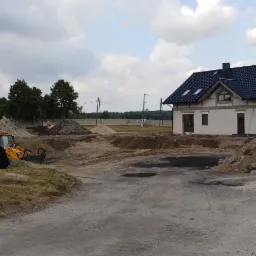 Żółta koparka i zielony teleskopowy podnośnik na placu budowy w Gryfinie, z nowym domem w tle i stertą gruzu na pierwszym planie.