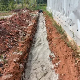 Wykop wypełniony piaskiem pod instalację sanitarną przy budynku z plandeką, widoczne fragmenty cegieł w wykopanej ziemi.