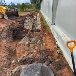 Tomo Bud - Wykopany rów wzdłuż białej hali namiotowej, widoczny żółty traktor z przodu, studzienka kanalizacyjna i łopata z pomarańczowym uchwytem, ziemia z gruzem ceglanym.