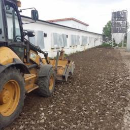 Tomo Bud - Żółta koparko-ładowarka wyrównuje teren podłoża z kamieni i ziemi przed budynkiem gospodarczym z silosami.