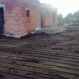 Surowy stan budynku z czerwonej cegły w trakcie budowy, widoczne fundamenty i przygotowany teren z równoległymi śladami po maszynach, prawdopodobnie pod przyszły trawnik lub kostkę.