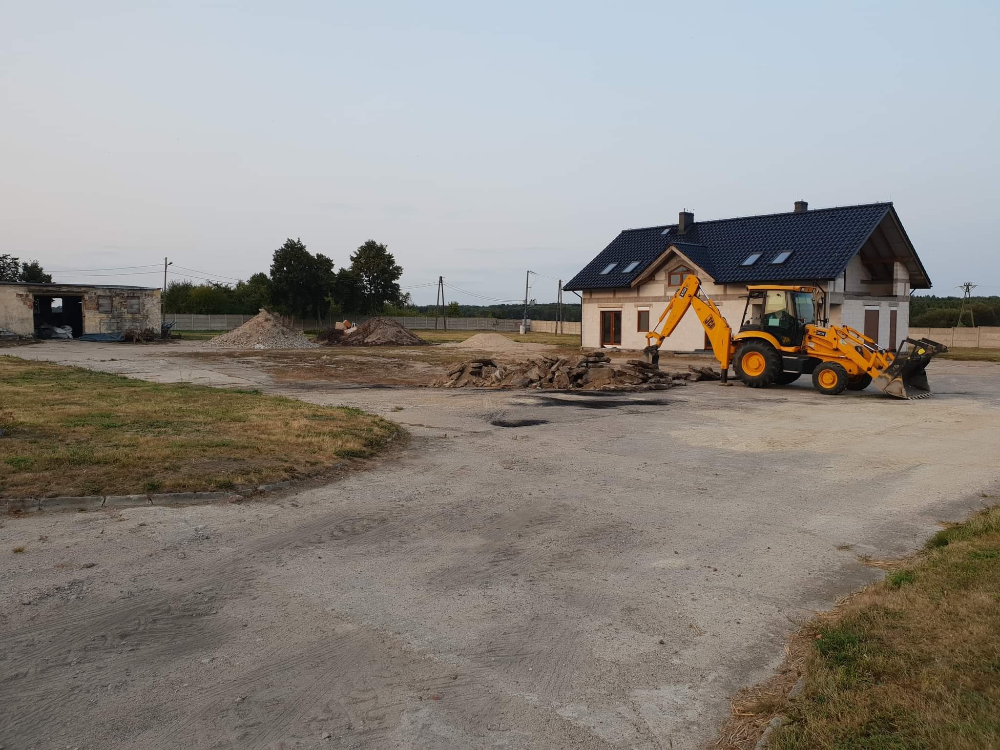 Żółta koparko-ładowarka JCB na placu budowy przed domem w stanie surowym otwartym; w tle stary, częściowo rozebrany budynek i hałdy ziemi