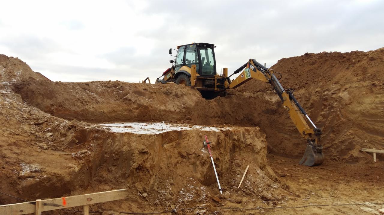 Żółta koparko-ładowarka CAT pracuje przy wykopie w gruncie gliniastym, widoczna łata białego śniegu, tyczka geodezyjna i drewniane szalunki.