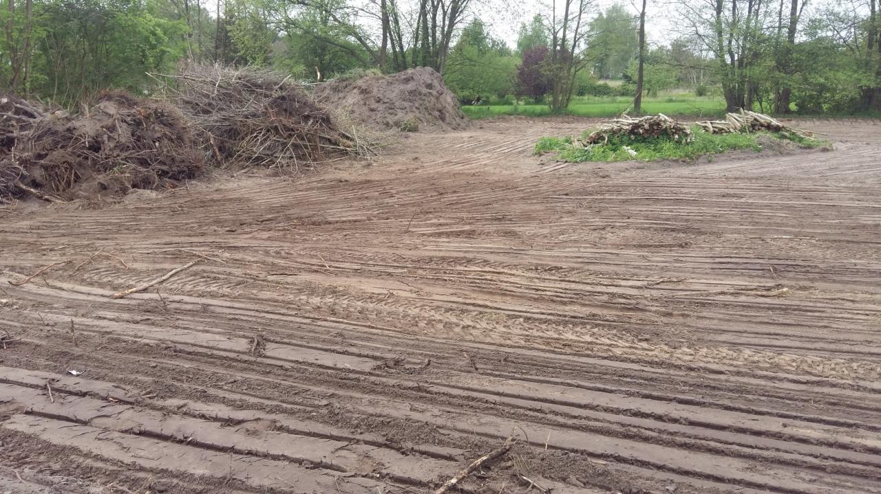 Przygotowany teren pod budowę z widocznymi śladami opon i stertami gałęzi oraz ziemi.