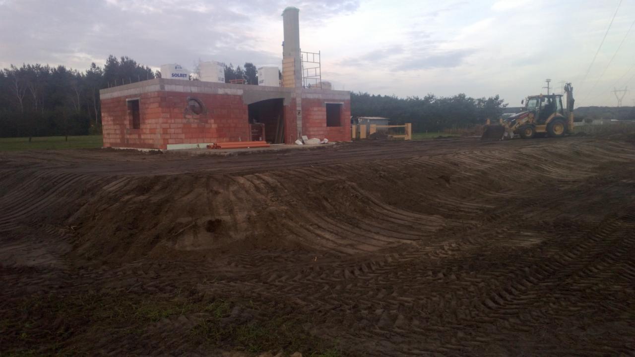 Nierówny teren po niwelacji z widocznymi śladami opon, w tle budynek w trakcie budowy z cegły, rusztowanie komina i koparko-ładowarka.