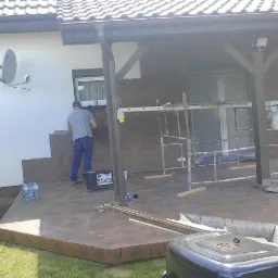 Montaż elewacji z cegły klinkierowej na budynku. Widoczne rusztowanie i narzędzia pracy. Prace w toku, częściowo ukończona ściana. Na ścianie antena satelitarna.