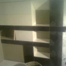 Wykończona ściana z płytek w łazience z wnękami, ciemne fugi i dekoracyjne wzory, ujęcie z dołu, widoczny fragment wiadra z zaprawą.