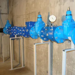 Błękitna instalacja wodna z zaworami i manometrem, wsparta na metalowych stelażach, na tle surowej, betonowej ściany z odręcznymi notatkami.