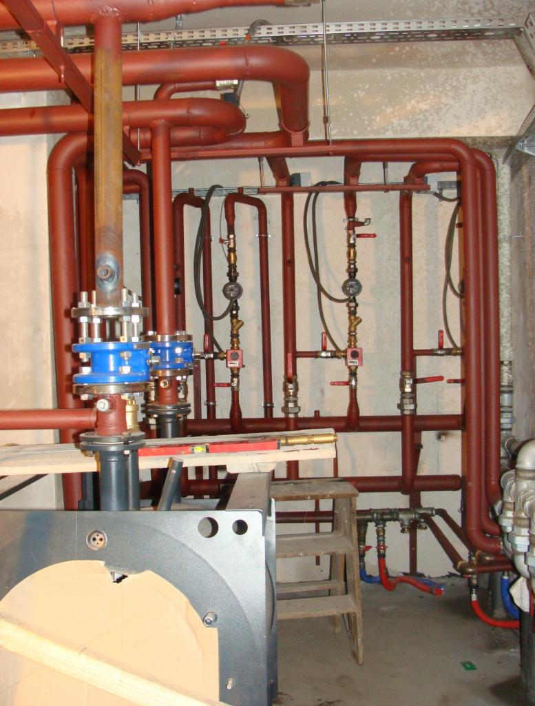 Rozbudowana instalacja grzewcza z czerwonymi rurami, zaworami i elementami montażowymi na tle betonowej ściany. Widoczny fragment pieca i poziomica.