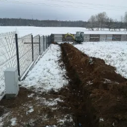Wykopany rów w zaśnieżonym terenie, obok ogrodzenia z siatki, z widoczną rurą i małą koparką w tle, sugerujący prace instalacyjne.