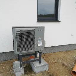 Szara pompa ciepła Nibe AMS10 zamontowana na betonowych fundamentach przy białej ścianie budynku z czarnym oknem, widoczny fragment rury kanalizacyjnej.