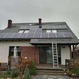 Dom jednorodzinny z zamontowanymi panelami fotowoltaicznymi na dachu pokrytym szarą dachówką, drabina oparta o elewację, jesienny ogród.