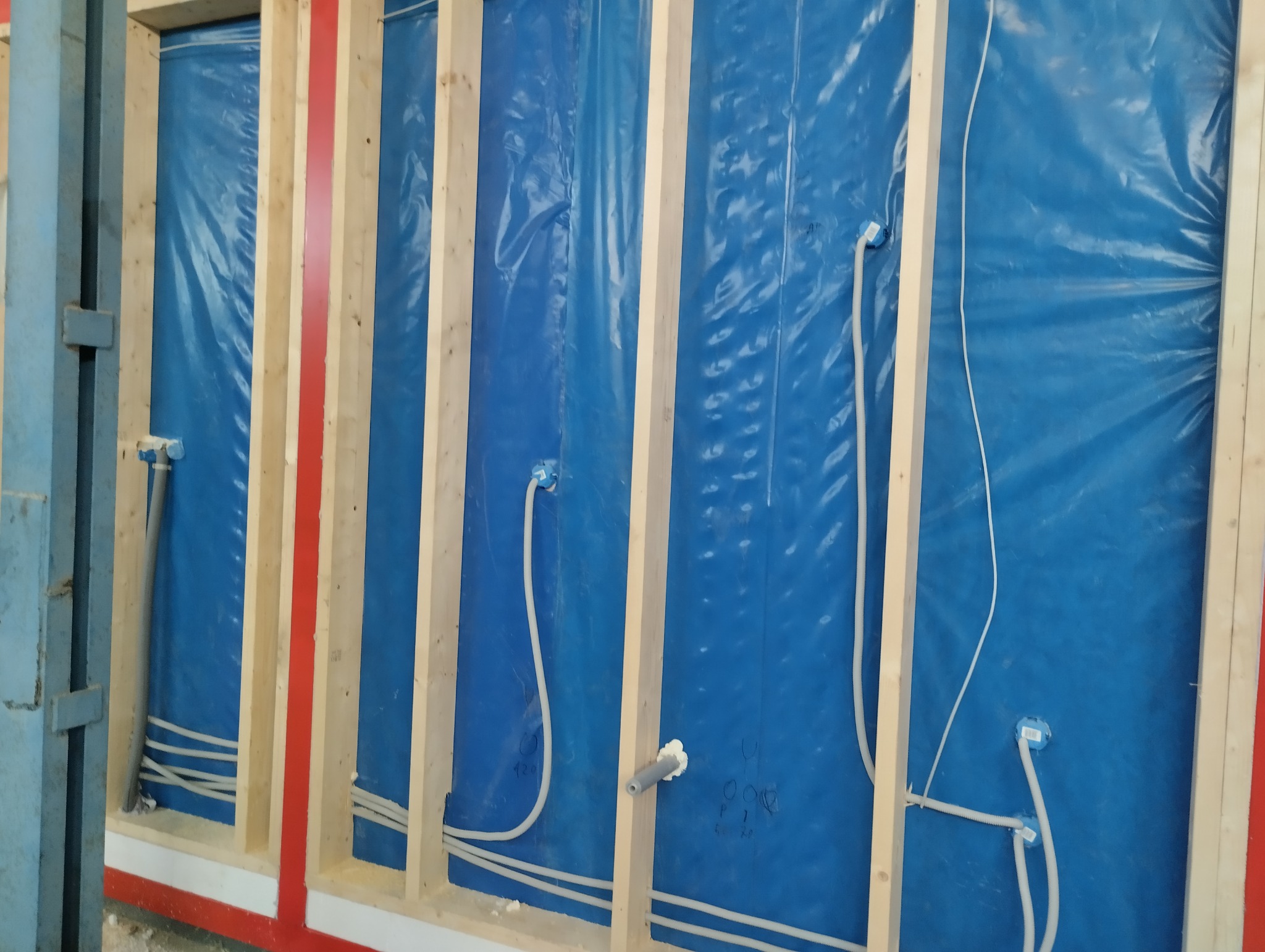 Ściana w trakcie budowy z widocznymi przewodami elektrycznymi i instalacją wodną, zabezpieczona niebieską folią, drewniane ramy konstrukcyjne.