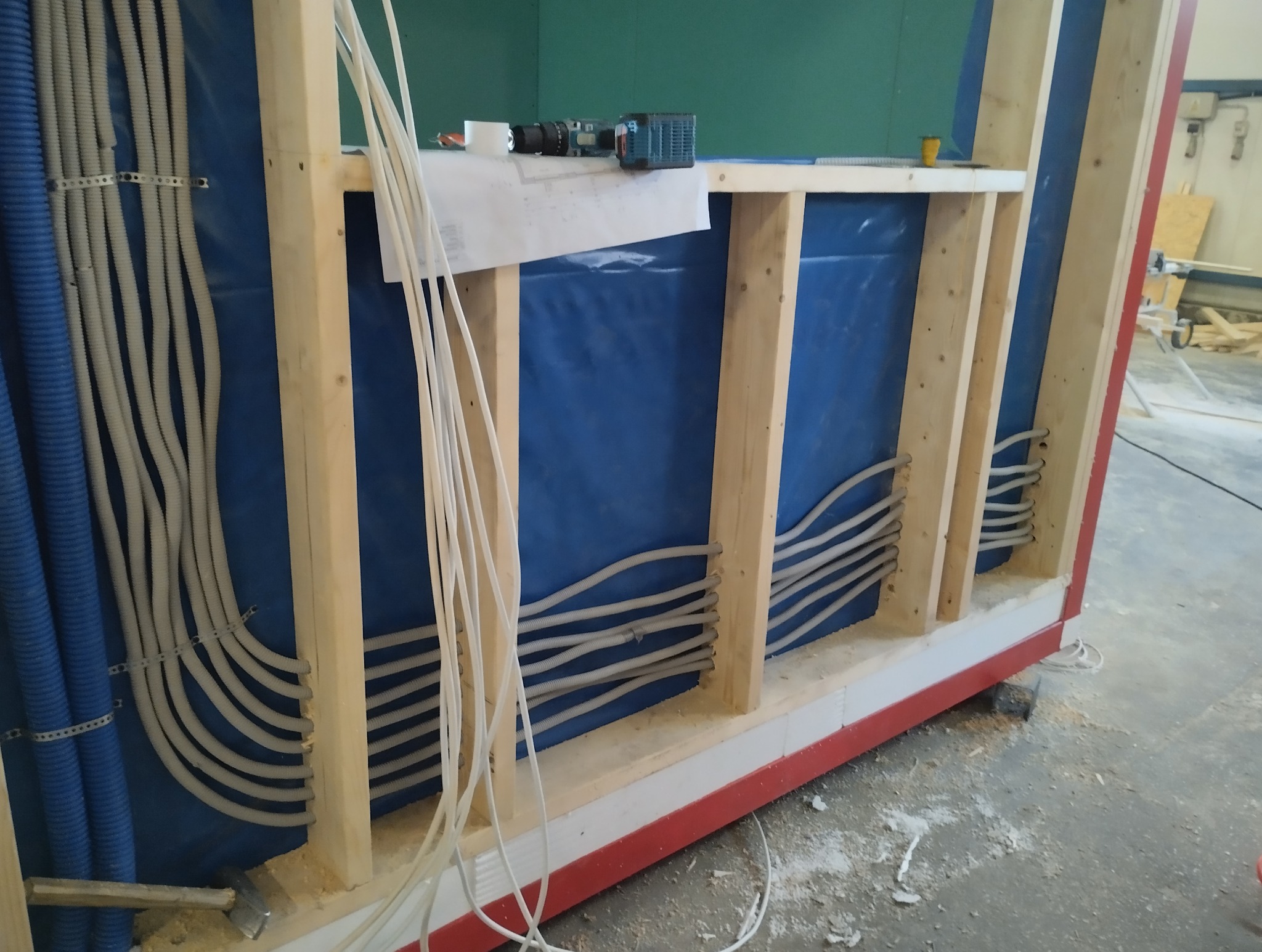 Wewnętrzna instalacja elektryczna w ścianie szkieletowej z niebieską folią, widoczne peszle z kablami, wkrętarka akumulatorowa na górnej belce konstrukcji.