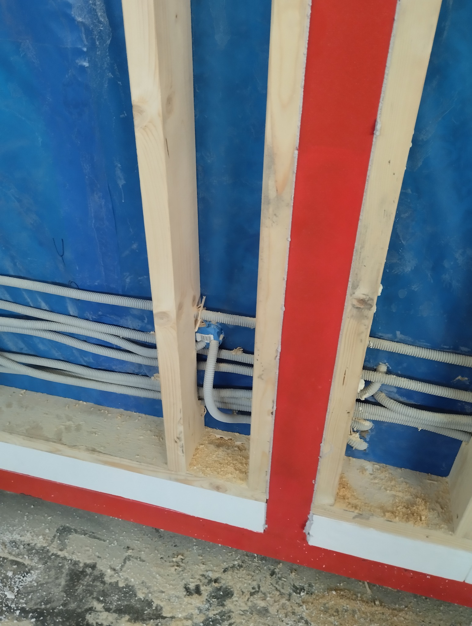 Wewnętrzna ściana szkieletowa w trakcie budowy z widocznymi peszlami instalacji elektrycznej, niebieską folią izolacyjną i elementami pomalowanymi na czerwono.