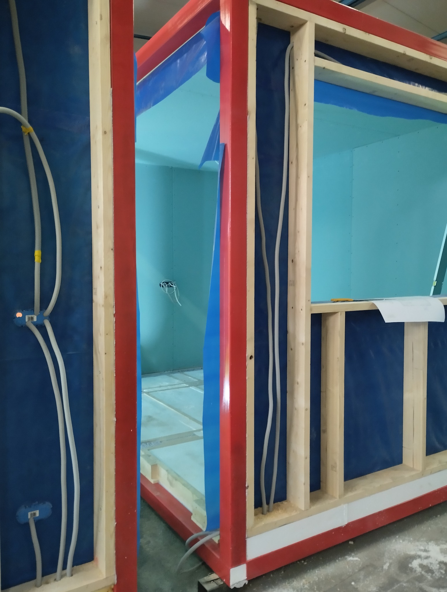 Wewnętrzna instalacja elektryczna w module kontenerowym, widoczne kable w peszlach prowadzonych w drewnianej konstrukcji szkieletowej, niebieska folia ochronna na ścianach.