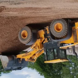 Żółta ładowarka Hyundai HL760-9 nabiera czerwonobrązowy materiał sypki, w tle widoczne bele słomy i drzewa.