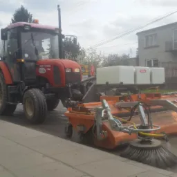 Czerwony ciągnik z pomarańczową szczotką czyszczącą pobocze drogi, w tle budynek z szyldem