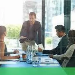 Mężczyzna w ciemnej marynarce prezentuje dokumenty grupie osób siedzących przy stole konferencyjnym w jasnym biurze; logo Schneider Electric w prawym górnym rogu.