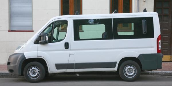 Biały bus pasażerski zaparkowany przed budynkiem z szarą roletą i brązowymi drzwiami.