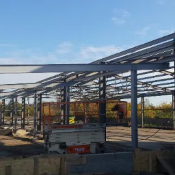 Konstrukcja stalowa hali w trakcie budowy, widoczny podnośnik koszowy i wykop pod fundamenty.