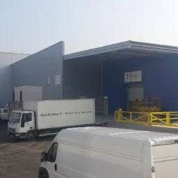 Magazyn z rampami załadunkowymi, widoczne białe busy i ciężarówki dostawcze na placu manewrowym.