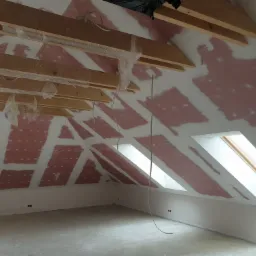 Poddasze w trakcie prac wykończeniowych: widoczne drewniane belki stropowe zabezpieczone folią, ściany ze śladami po szpachlowaniu, dwa okna dachowe w skosie.