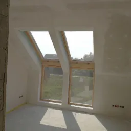 Poddasze ze skośnym sufitem, dwa okna dachowe z drewnianymi ramami, surowe ściany po szpachlowaniu, widoczne puszki elektryczne i żółta taśma malarska przy podłodze.