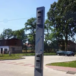 Słupkowy domofon wielorodzinny z klawiaturą numeryczną i głośnikiem, zamontowany na zewnątrz budynku, z widoczną folią ochronną na obudowie, na tle wiejskiej zabudowy i samochodów.