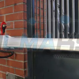 Zamontowany napęd bramy skrzydłowej z centralą sterującą na ceglanym słupku, widoczna lampa ostrzegawcza i fragment metalowej bramy.