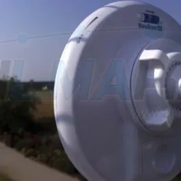 Biała antena NanoBeam zamocowana na metalowym słupie z widocznymi śrubami mocującymi, w tle rozmyty krajobraz wiejski pod błękitnym niebem.