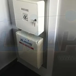 Biała szafka na klucz do wentylacji pożarowej z symbolem płomienia i kluczem w zamku, zamontowana nad zwiniętą drabinką ewakuacyjną z logo firmy ZILMAKS i numerem telefonu na białej ścianie.