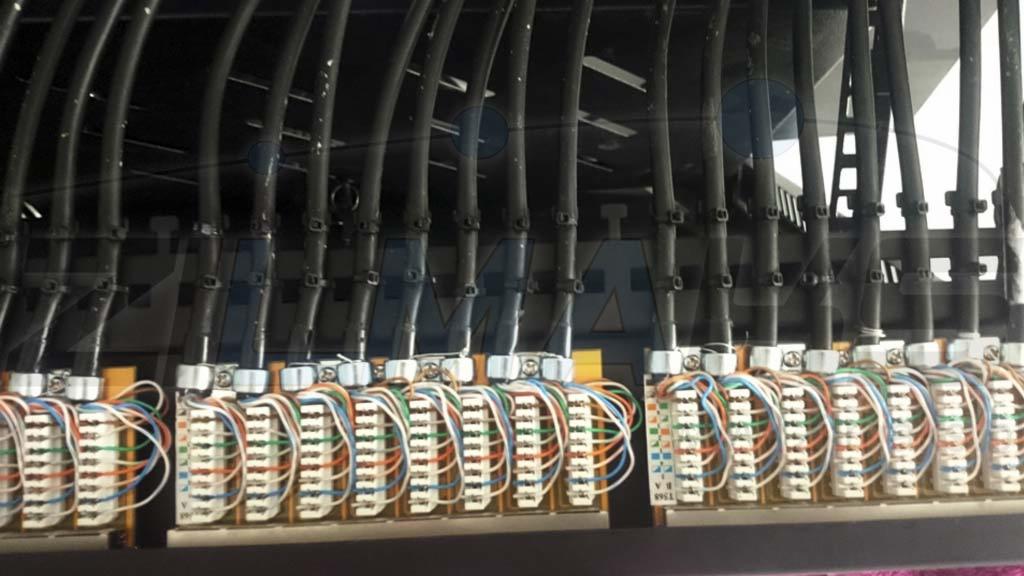 Rząd złączek kablowych z podłączonymi przewodami w kolorach: białym, niebieskim, pomarańczowym i zielonym. Kable prowadzone w czarnych peszlach, przymocowanych do konstrukcji. Widoczne oznaczenia...