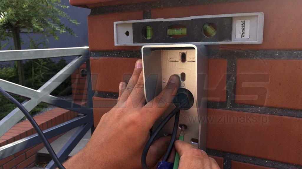 Instalacja wideodomofonu: monter podłącza przewody w puszce montażowej na ceglanym słupku ogrodzenia, poziomica Makita na górze, widoczne narzędzia.