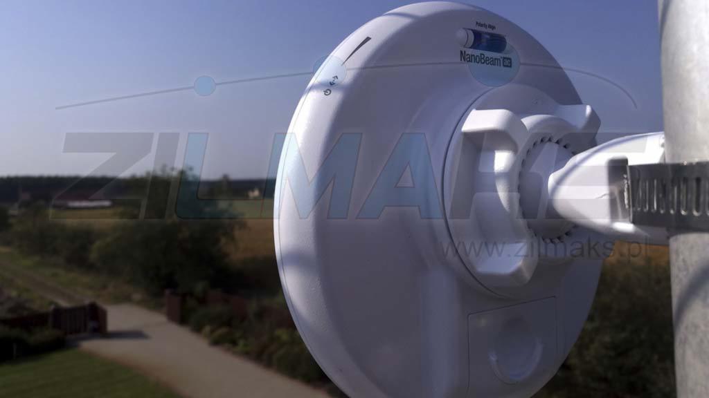 Biała antena NanoBeam zamocowana na metalowym słupie z widocznymi śrubami mocującymi, w tle rozmyty krajobraz wiejski pod błękitnym niebem.